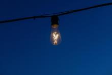 Lamparilla eléctrica encendida, conectada con un cable a ambos lados, a la que se le puede ver el filamento. Fondo azul oscuro. 