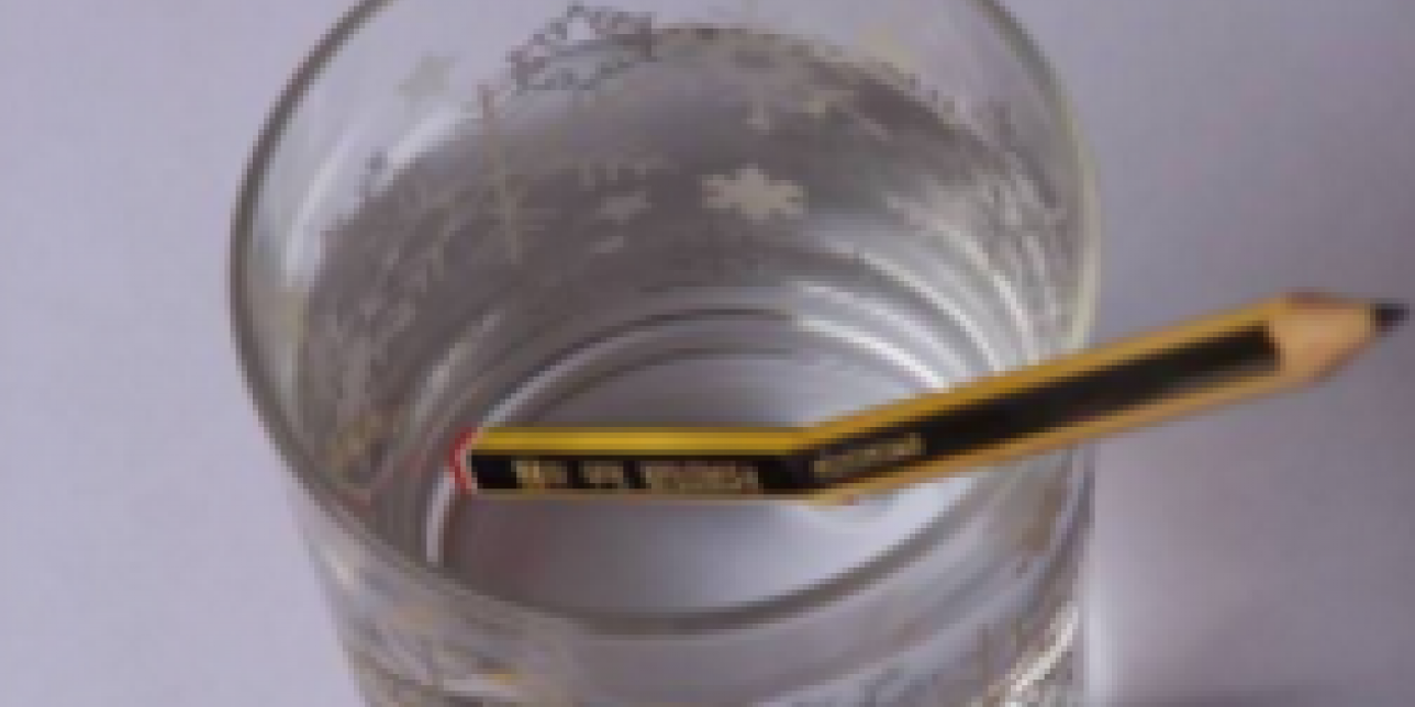 Vaso con agua y un lápiz amarillo y negro. La mitad del lápiz se encuentra sumergido en el agua y la foto es tomada desde arriba. Se observa el fenómeno de refracción.