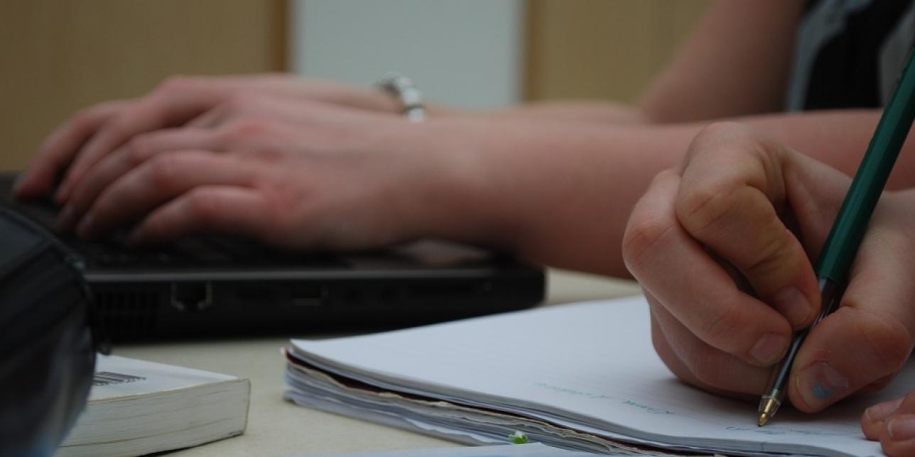 Imagen que muestra las manos de dos personas adultas en actitud de aprendizaje (escribiendo sobre un papel y apoyándose sobre un ordenador)