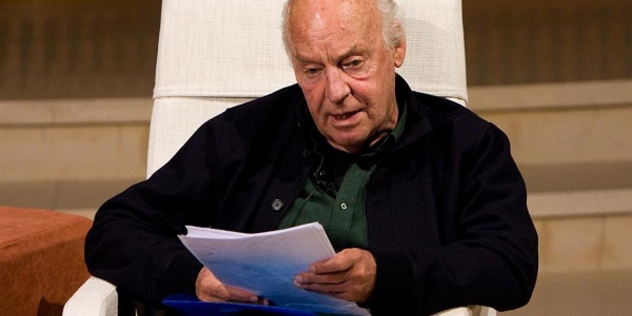 Fotografía de Eduardo Galeano en plano medio, sentado en un sofá y con unas hojas en su mano.
