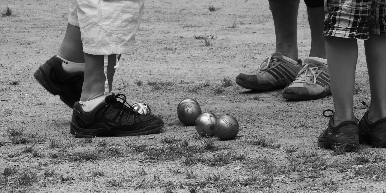 Foto en blanco y negro con pies de niños, con zapatos, entorno a unas bochas sobre el suelo de tierra
