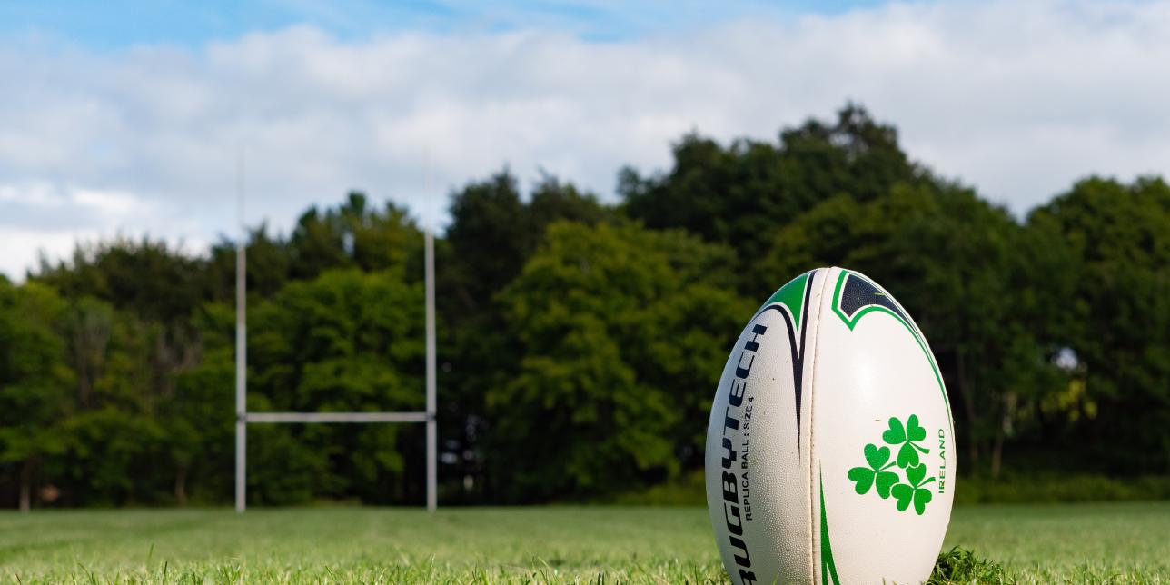 Imagen decorativa de una cancha de rugby con un primerisimo primer plano de una pelota y en el fondo la H o portería de rugby