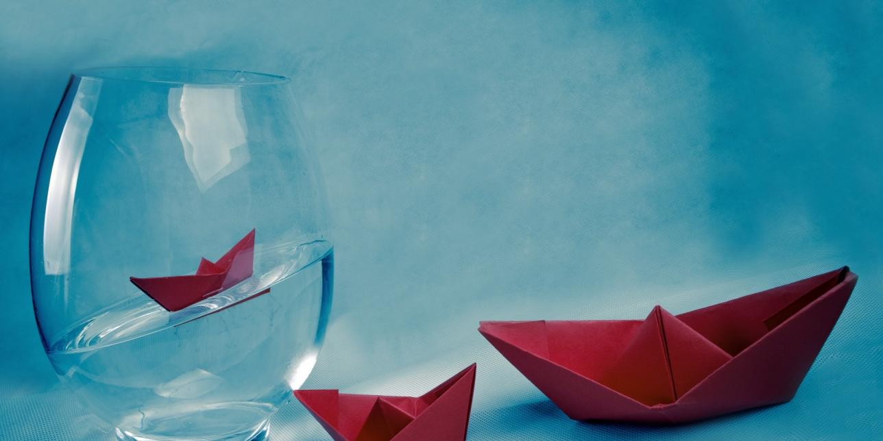 Vaso de vidrio con agua y tres barcos de papel plegado.