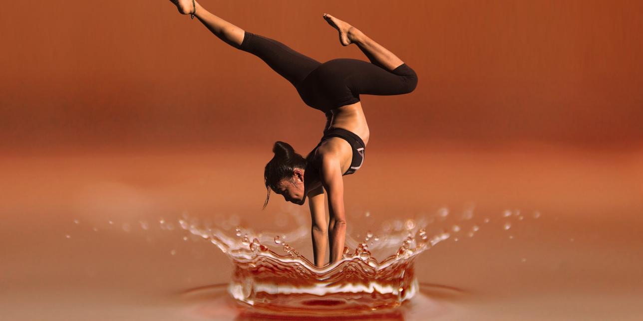 Mujer realizando una parada de manos sobre una superficie que simula agua, con un fondo degradado en tonos marrones. La postura refleja equilibrio y fuerza, mientras el agua alrededor forma un efecto visual de salpicadura.